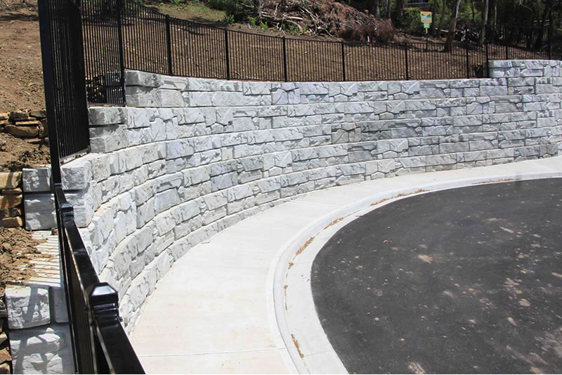 Curved gray stone retaining wall alongside a sidewalk and asphalt road, bordered by black metal fencing.