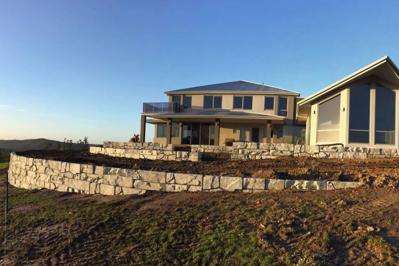 Modern two-story house with large windows and multiple terraced retaining walls made of stone blocks, under a clear blue sky at sunset.