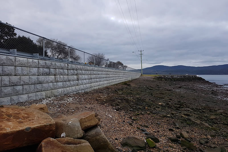 Stone retaining wall with metal fencing along a rocky shoreline under a cloudy sky.