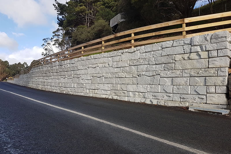 Stone retaining wall with a wooden railing along a paved road bordered by trees.