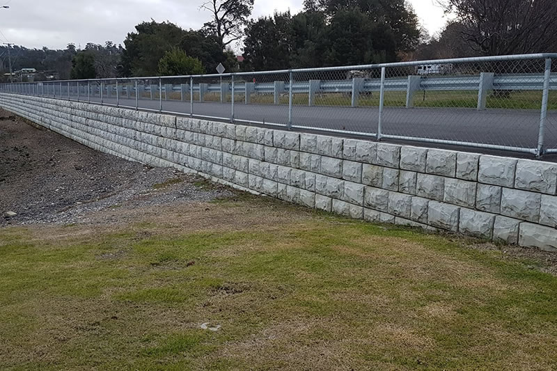 Long stone retaining wall with a metal fence on top, adjacent to a grassy area and a paved road, under a cloudy sky.