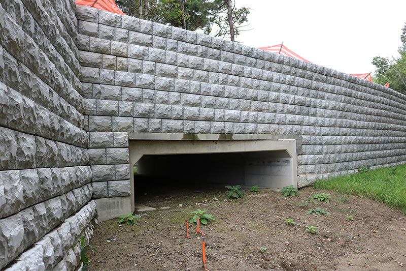 Large stone retaining wall with rectangular tunnel opening at the base, surrounded by dirt and sparse vegetation.