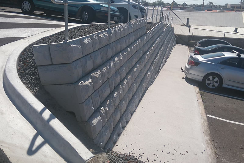 Concrete retaining wall with textured blocks supporting a landscaped area next to a parking lot with parked cars.