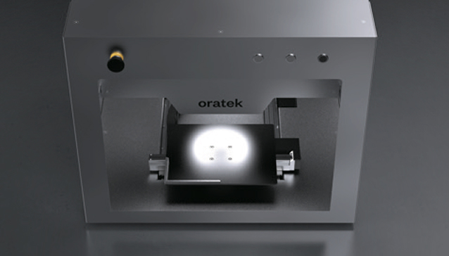 Close-up of a metallic device with the brand name oratek and a lit platform inside.
