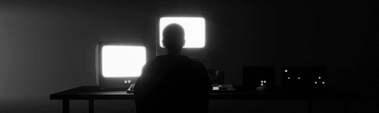 Silhouette of a person sitting at a desk with two glowing computer monitors in a dark room.