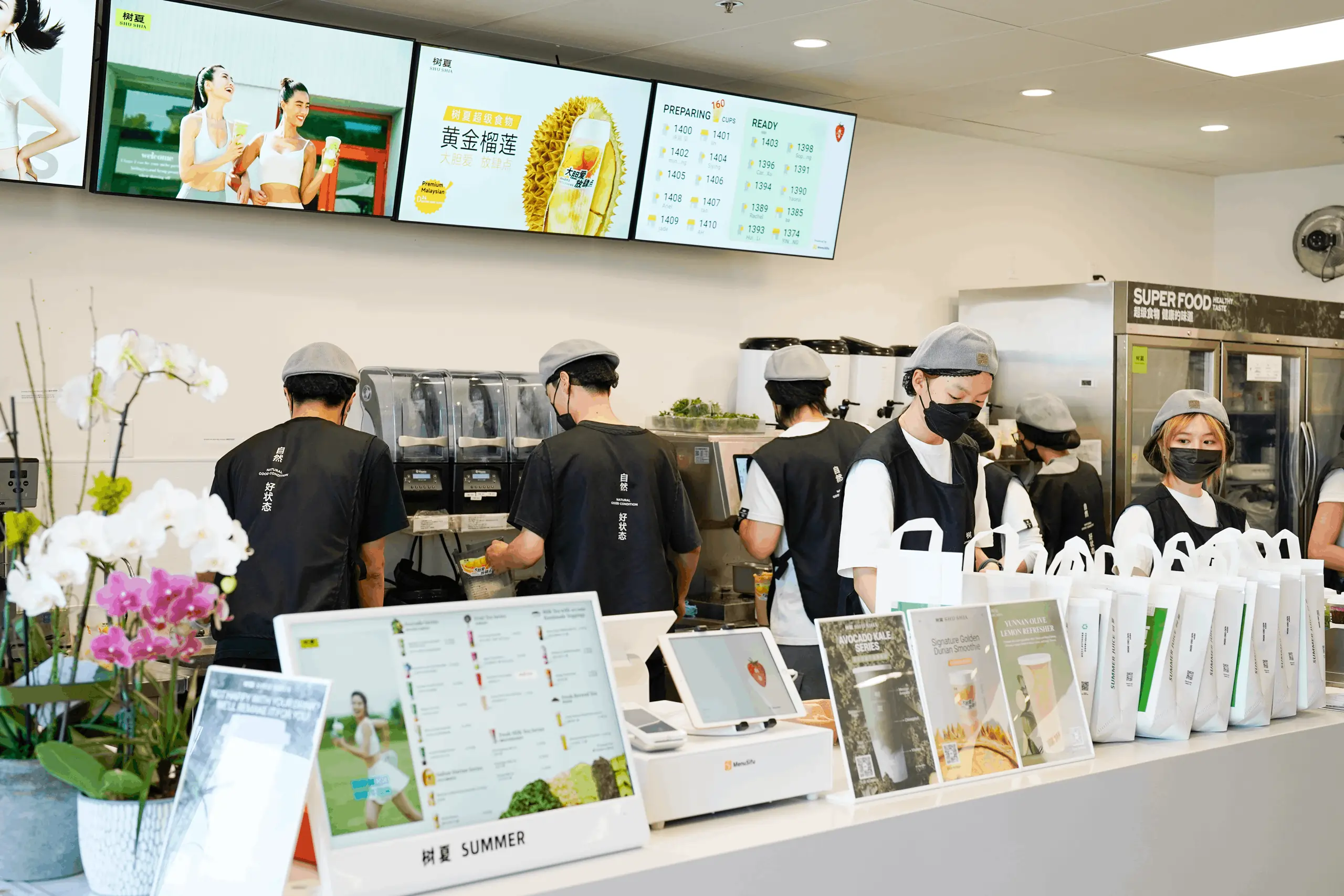 Busy staff in modern bubble tea shop assemble drinks under an order status screen showing real-time updates for customer orders.