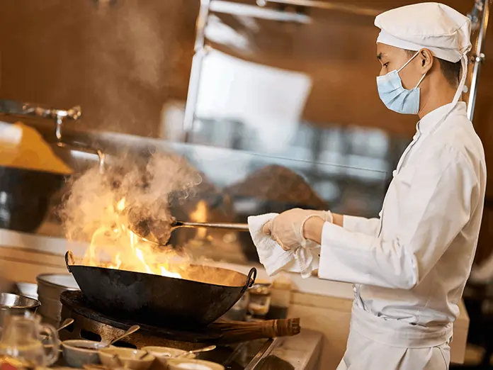 Chef wearing mask and uniform cooks with high flames in a busy kitchen, managing orders through a kitchen display system.