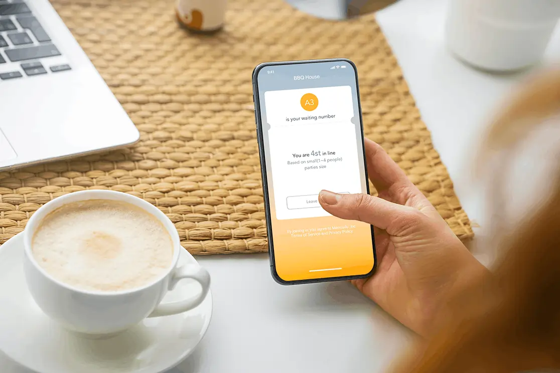 Customer checks restaurant waitlist app on smartphone to track queue position while enjoying coffee at a cafe table.
