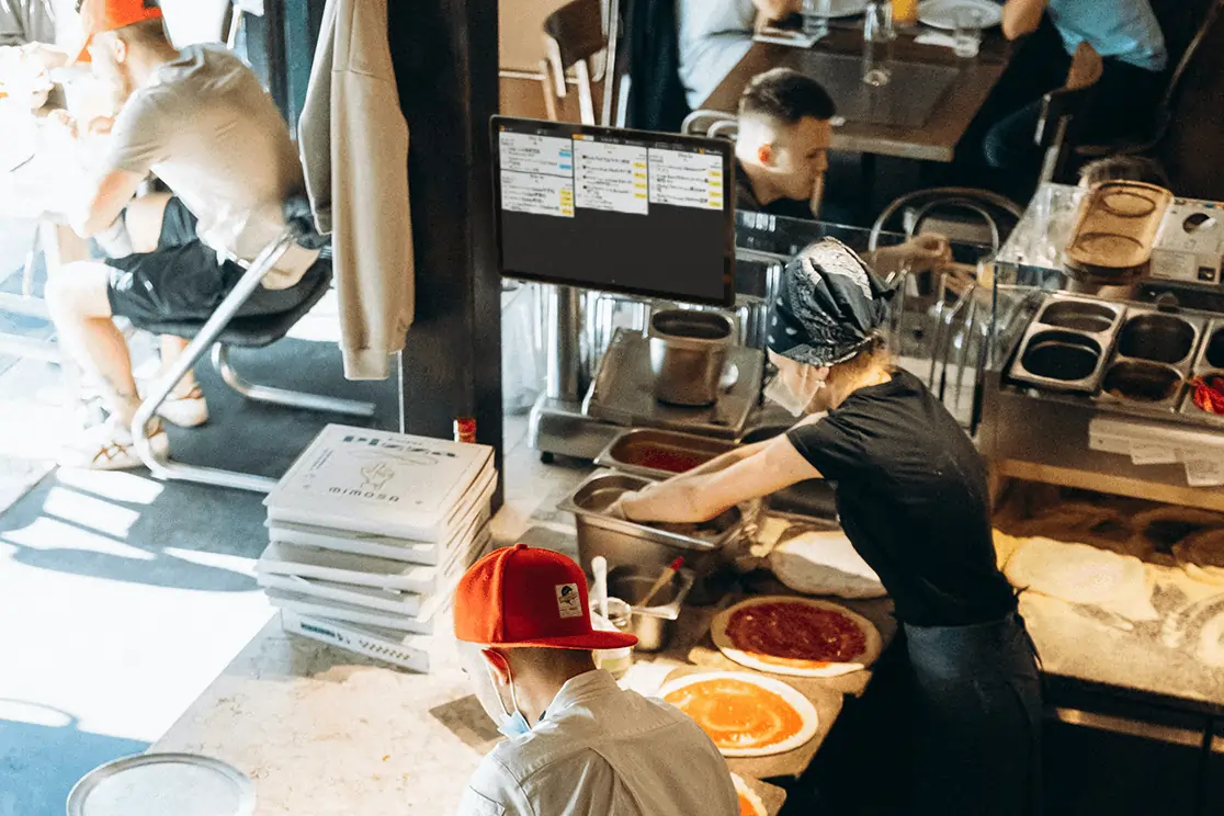Kitchen staff assembling pizzas with real-time orders shown on a KDS screen to streamline workflow and speed service.