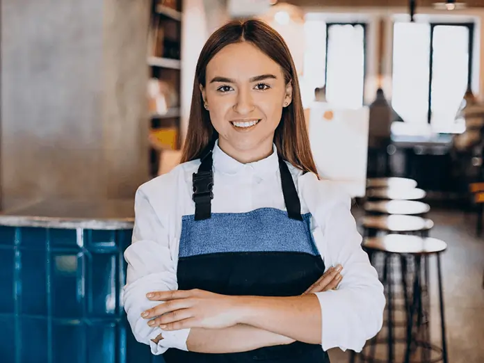 Smiling restaurant staff member in apron standing confidently with arms crossed inside a bright, modern restaurant.