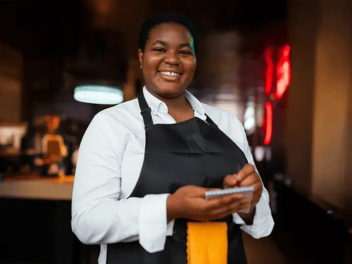 Server in crisp white shirt and dark apron smiles while holding a pen and order pad in a softly lit restaurant interior.