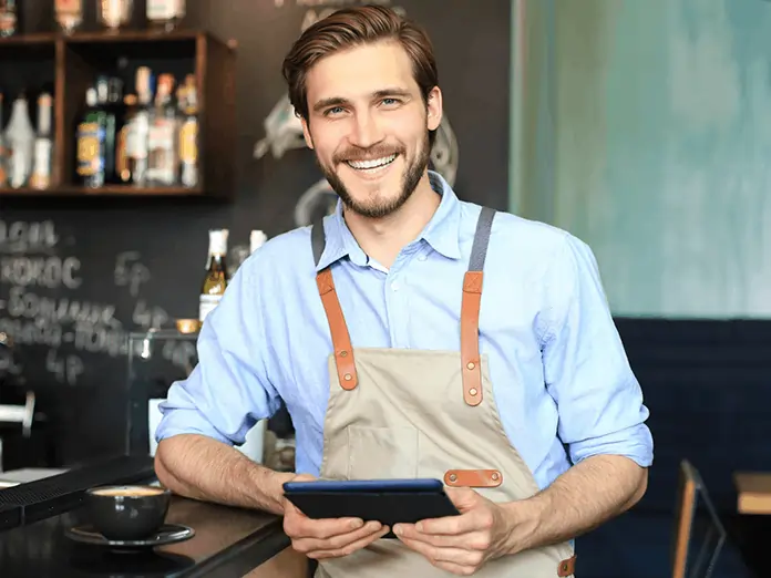 Smiling café employee in a blue shirt and apron holds a tablet while standing behind the counter in a cozy coffee shop.