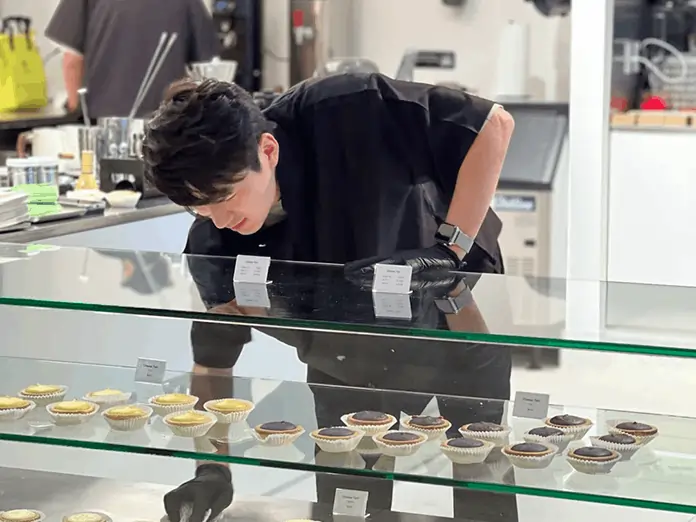 A staff member arranges tarts in a bakery display case at Kyo Mart, featured in bakery cafe POS system testimonial image.