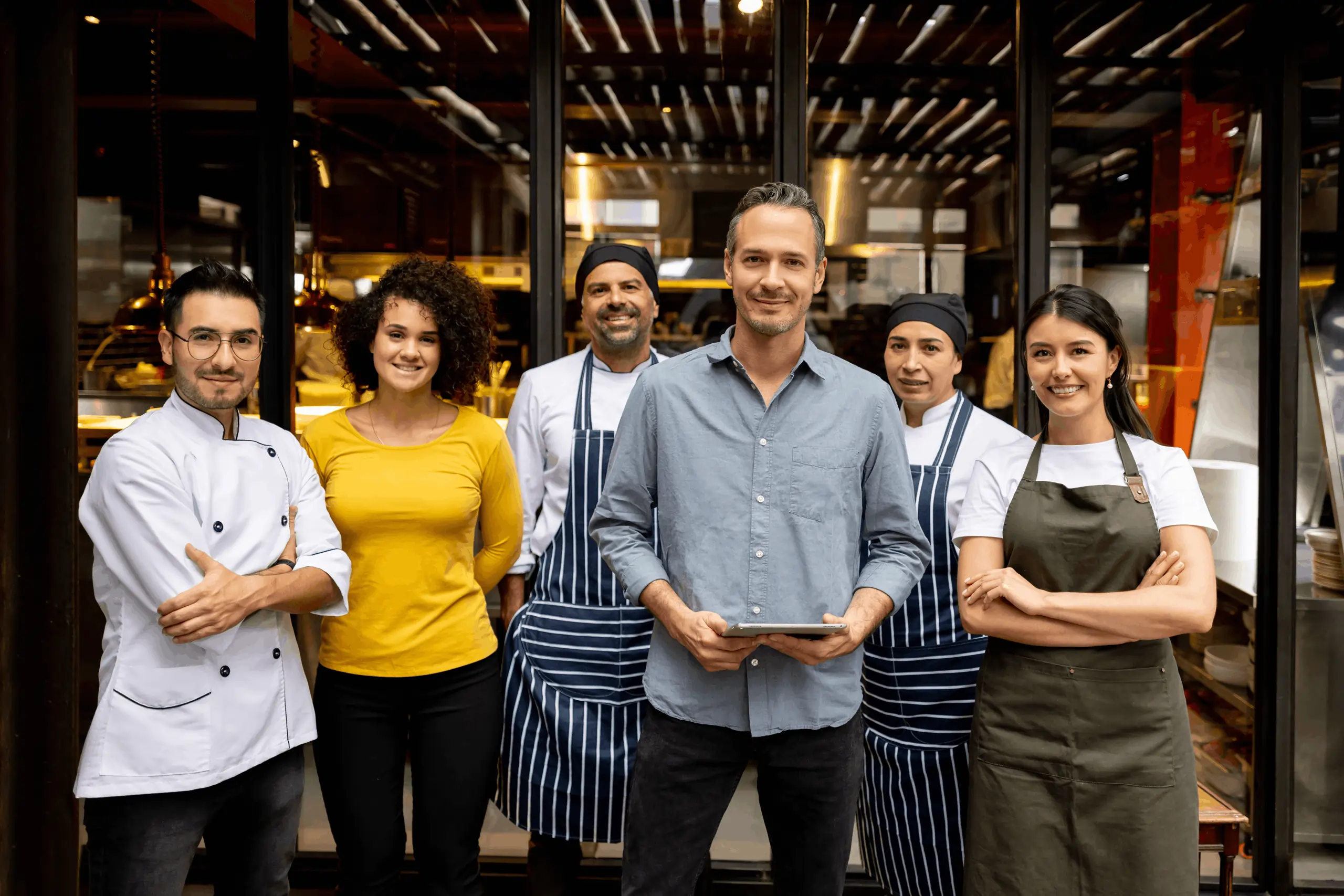 Smiling restaurant crew including chefs, servers, and owner posing confidently in front of the kitchen entrance.