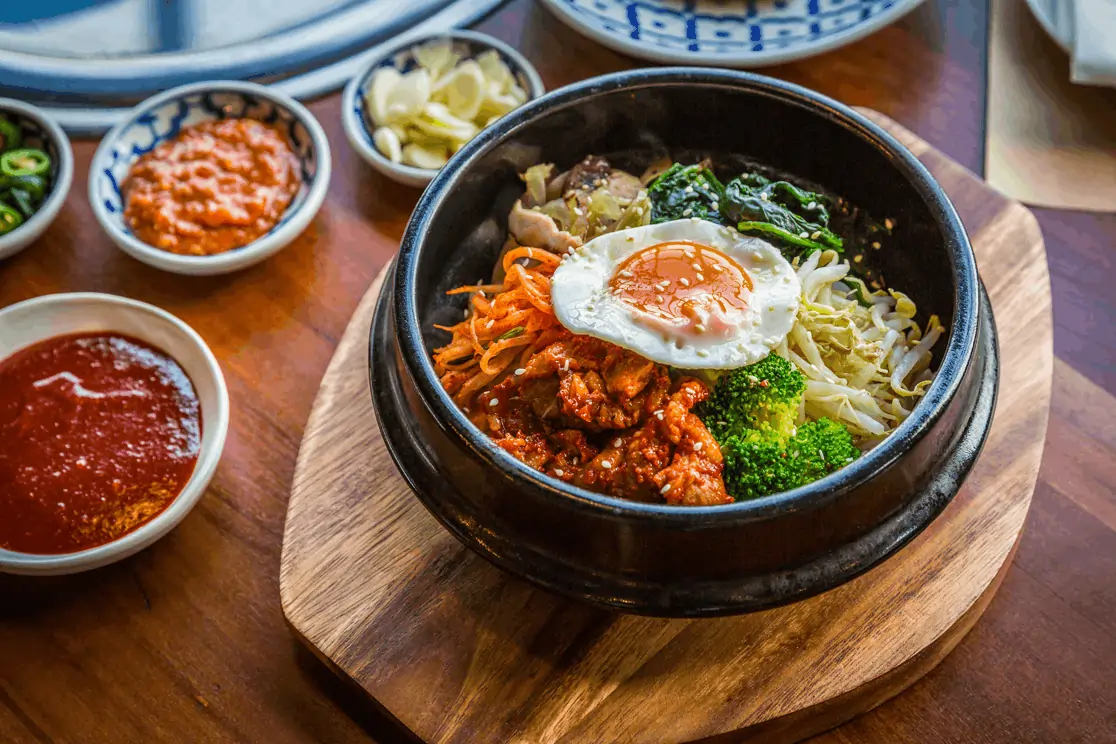 Traditional Korean bibimbap in a sizzling stone bowl topped with egg and surrounded by colorful side dishes on a wooden table.