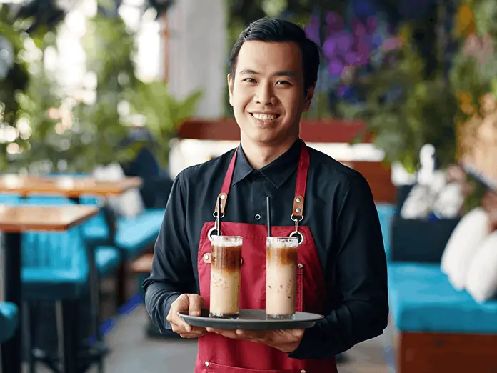 Smiling Vietnamese café server holds two iced coffees, praising the speed and ease of the Vietnamese POS system at work.