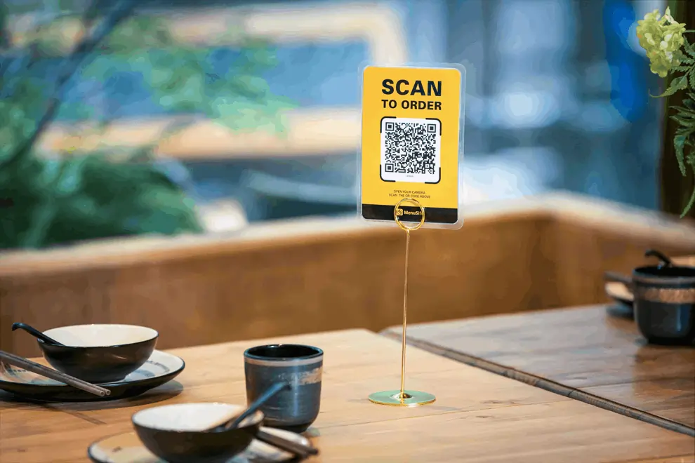 A yellow tabletop sign with a QR code reads “Scan to Order,” placed on a wooden table in a restaurant with bowls and chopsticks.