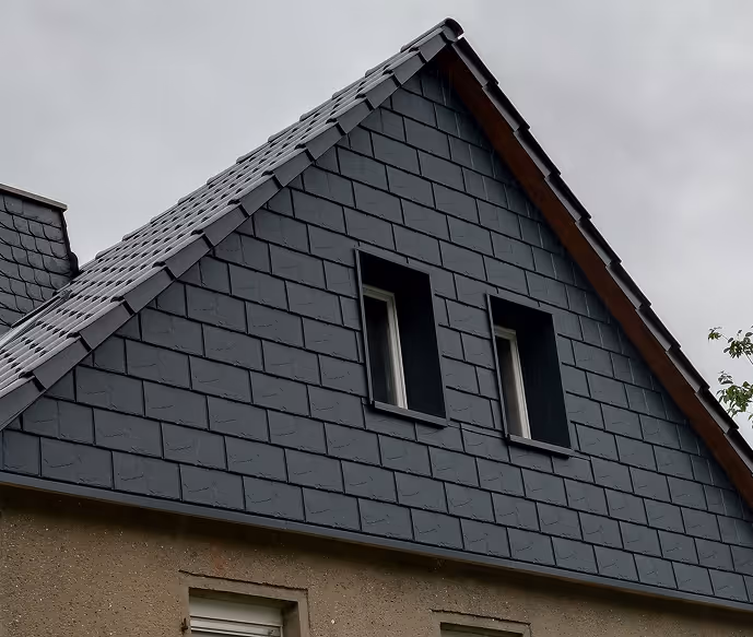 Spitzdach eines Hauses mit dunkelgrauen Schieferplatten und zwei schmalen Fenstern bei bewölktem Himmel.