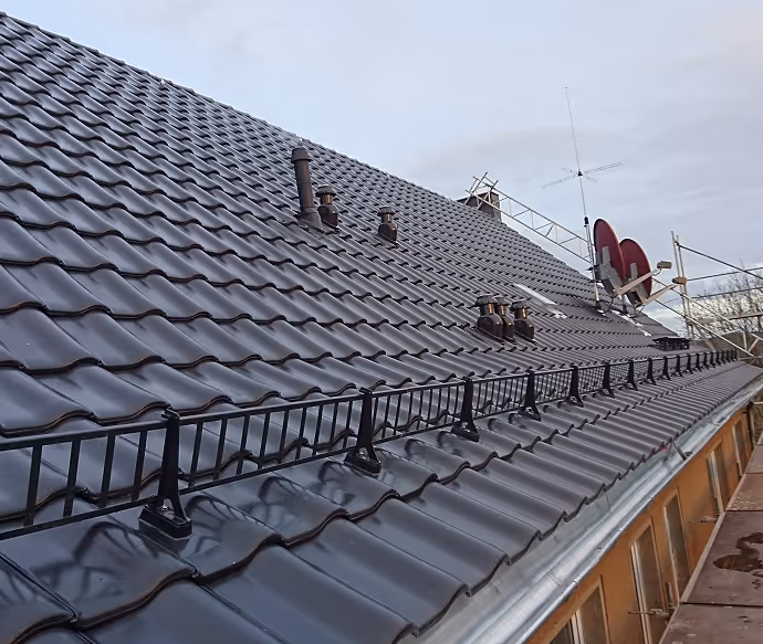 Detailaufnahme eines nassen schwarzen Ziegeldachs mit Schornsteinrohren, Antennen und Satellitenschüsseln vor bewölktem Himmel.