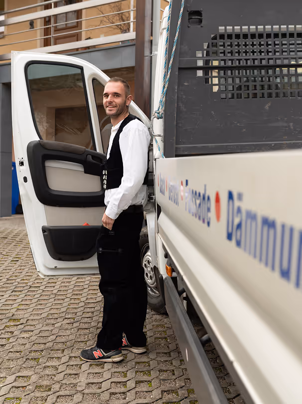 Ein lächelnder Mann in traditioneller Handwerkskleidung steht neben einem weißen Transporter mit geöffneter Fahrertür.
