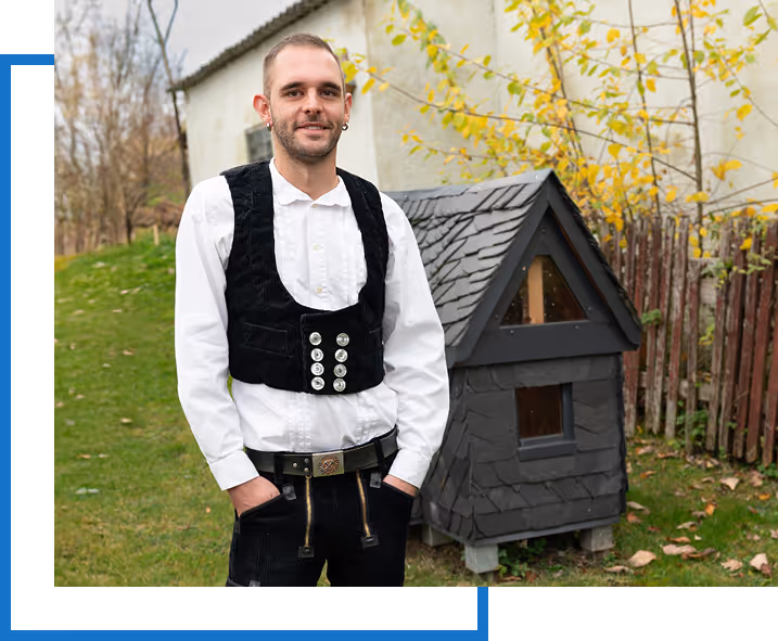 Ein Mann in traditioneller schwarzer Arbeitskleidung mit bestickter Weste und weißem Hemd steht im Garten vor einem kleinen schwarzen Hundehäuschen.