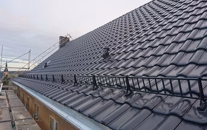 Dach mit schwarzen glänzenden Ziegeln und installiertem Schneefanggitter unter bewölktem Himmel.