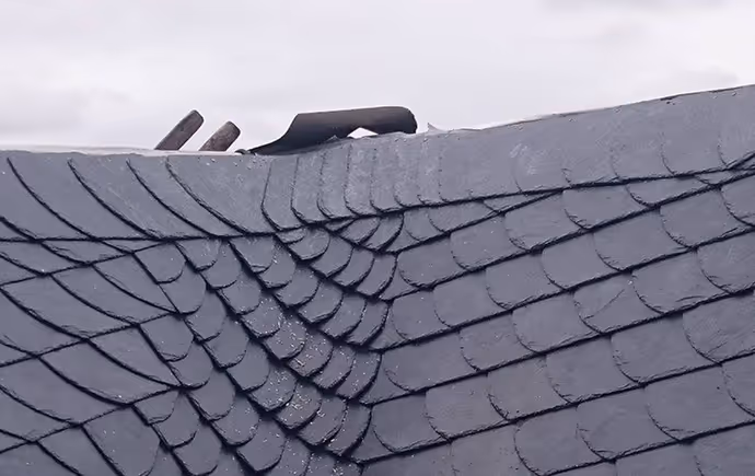 Nahaufnahme von einem Dach mit kunstvoll angeordneten grauen Schieferziegeln und einem Teil eines Schornsteins.