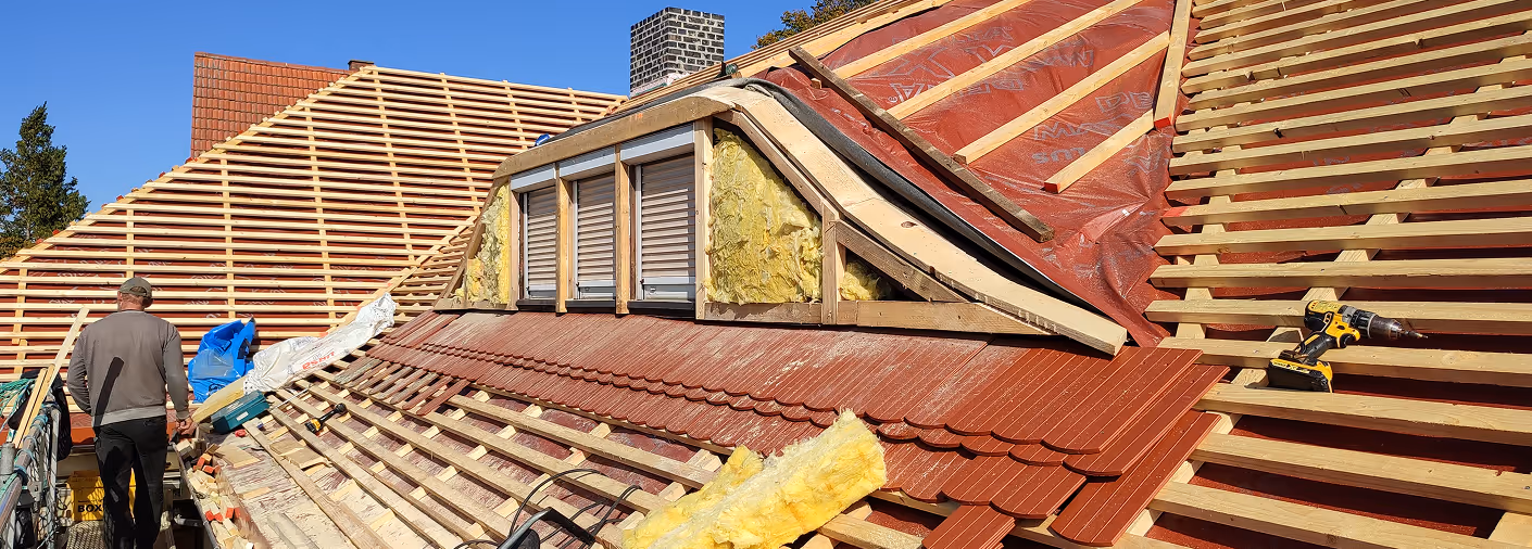 Eine Baustelle auf einem Hausdach mit teilweise verlegten roten Dachziegeln, Dämmmaterial und einem Arbeiter auf einem Gerüst.