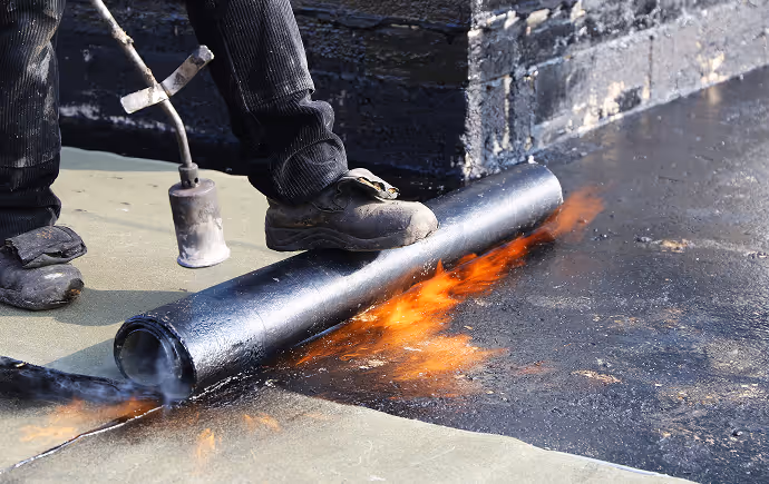 Person applying fire to roll of waterproofing membrane on rooftop for roofing work.