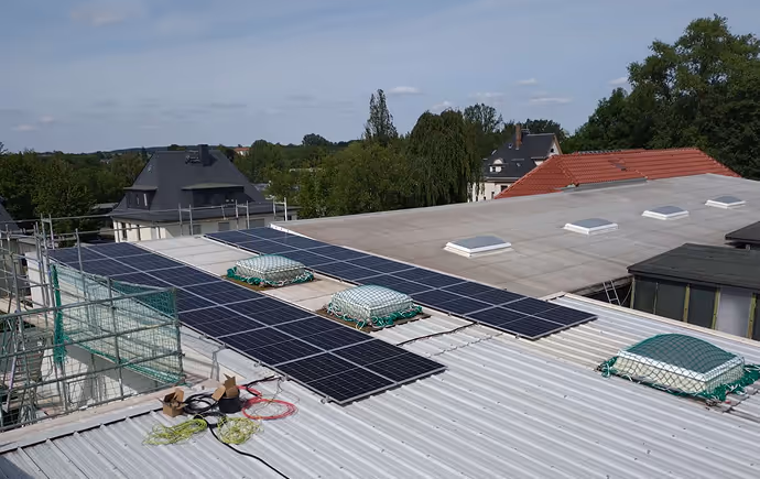 Auf einem flachen Dach sind mehrere Solarmodule installiert, umgeben von einem Gerüst und mit grünem Umfeld im Hintergrund.