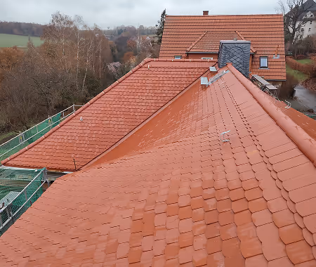 Neues, rot gekacheltes Dach eines Hauses mit Blick auf benachbarte Dächer und bewaldete Landschaft.