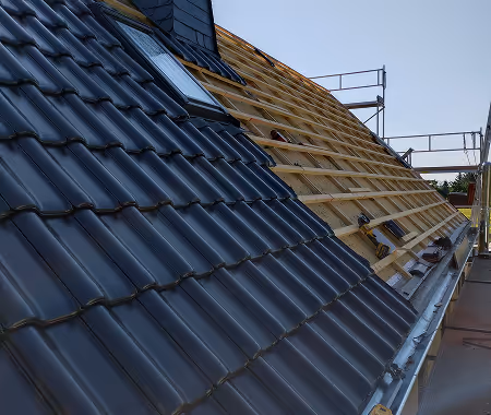 Teilweise mit schwarzen Dachziegeln eingedecktes Schrägdach neben unbedecktem Holzgerüst, mit Gerüst im Hintergrund.