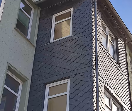 Fassade eines Gebäudes mit blaugrauen, schuppenförmigen Verkleidungsplatten und weißen Fenstern.