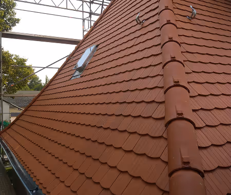 Schrägdach mit roten Tonziegeln und kleiner Dachbodenluke unter einem Gerüst.