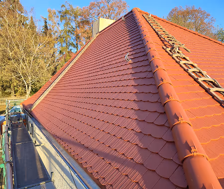 Rotes Ziegeldach eines Hauses bei sonnigem Wetter mit Bäumen im Hintergrund.