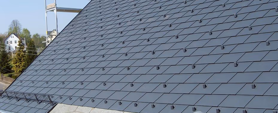 Dach mit grauen Schieferziegeln und Schneefanghaken, Teil eines Wohnhauses im Hintergrund sichtbar.