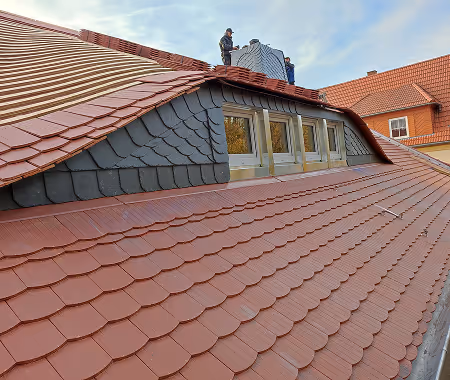 Zwei Handwerker verlegen Dachziegel auf einem Dach mit Gaubenfenstern bei klarem Himmel.