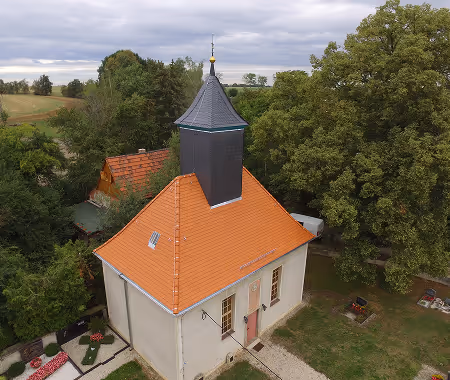 Luftaufnahme einer kleinen Kirche mit rotem Ziegeldach und dunklem Turm, umgeben von grünen Bäumen und Feldern unter bewölktem Himmel.