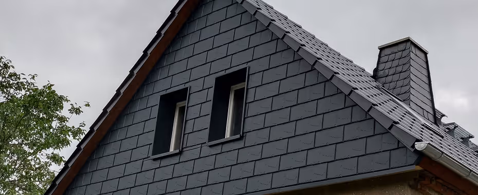 Giebel eines Hauses mit grauem Schieferdach und zwei kleinen Fenstern unter bewölktem Himmel.