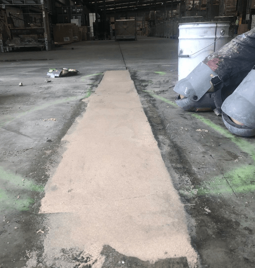 Fresh concrete patch repair applied on a warehouse floor with a worker’s feet and a white bucket nearby.
