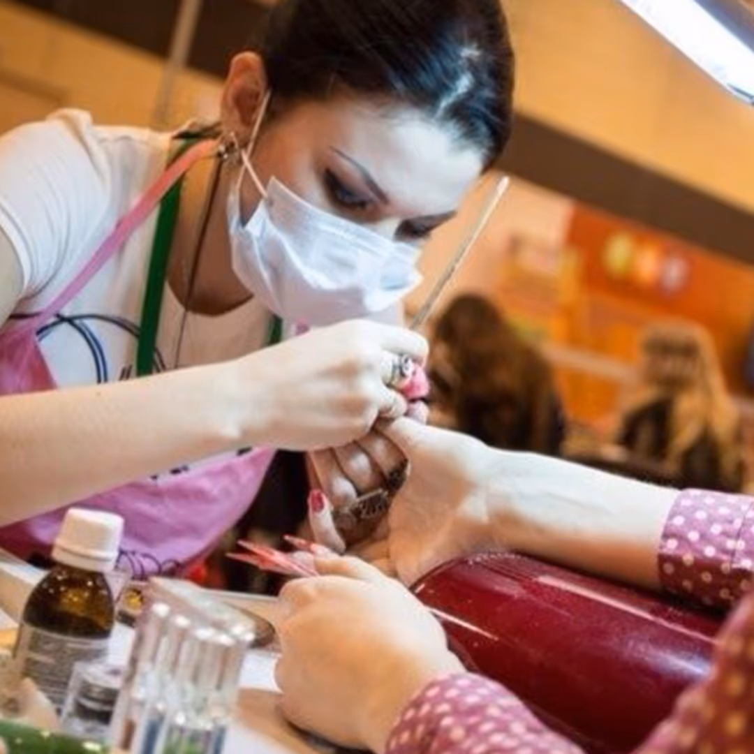 April Ryan wearing mask giving a manicure to a client in a salon with nail care products nearby.