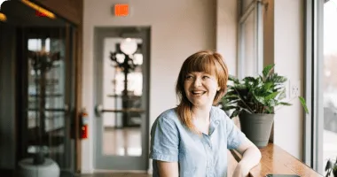 Woman with red hair smiling while sitting by a window with a potted plant in a cozy room.