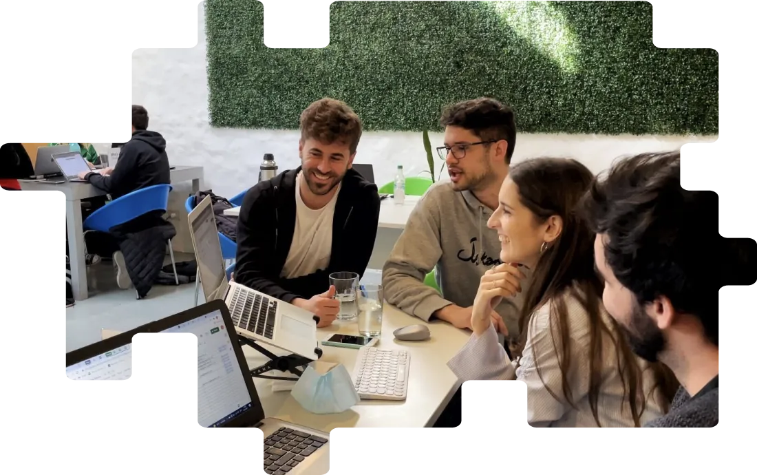 Four young adults sitting around a table in a modern office, smiling and collaborating with laptops and a glass of water on the table.