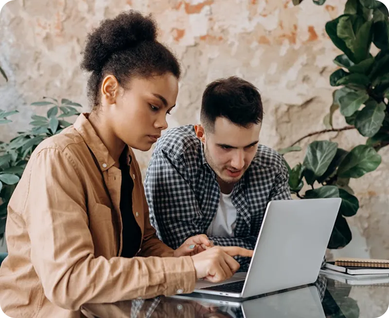 Two people, a woman and a man, focused on a laptop screen while working together at a glass table indoors.
