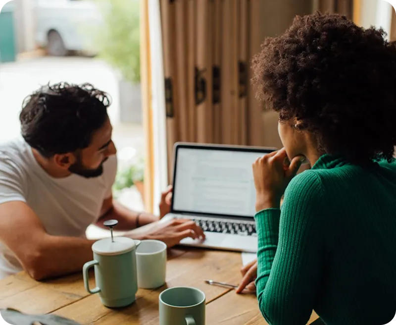 Man and woman sitting at a table with coffee cups, looking at a laptop screen together.