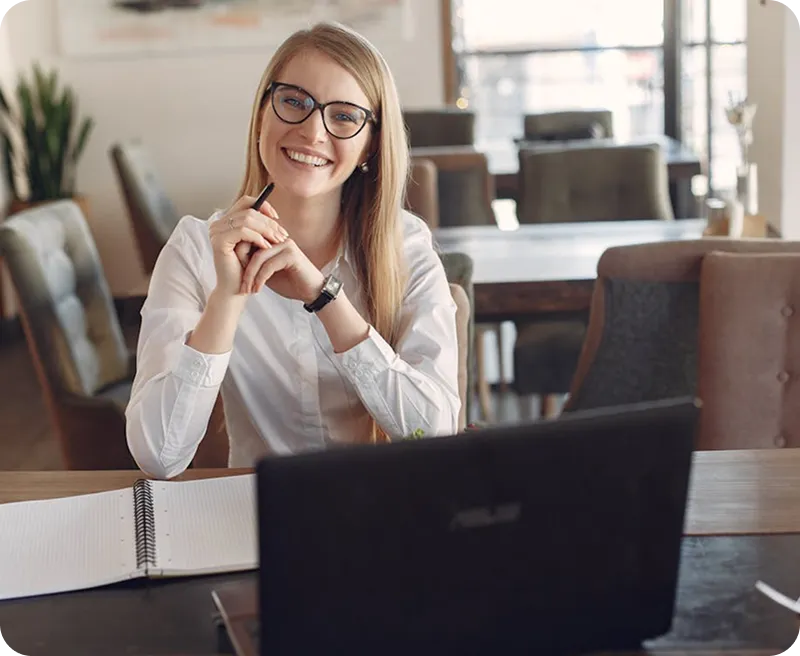 Smiling woman with glasses sitting at a table with a notebook and laptop in a bright office setting.