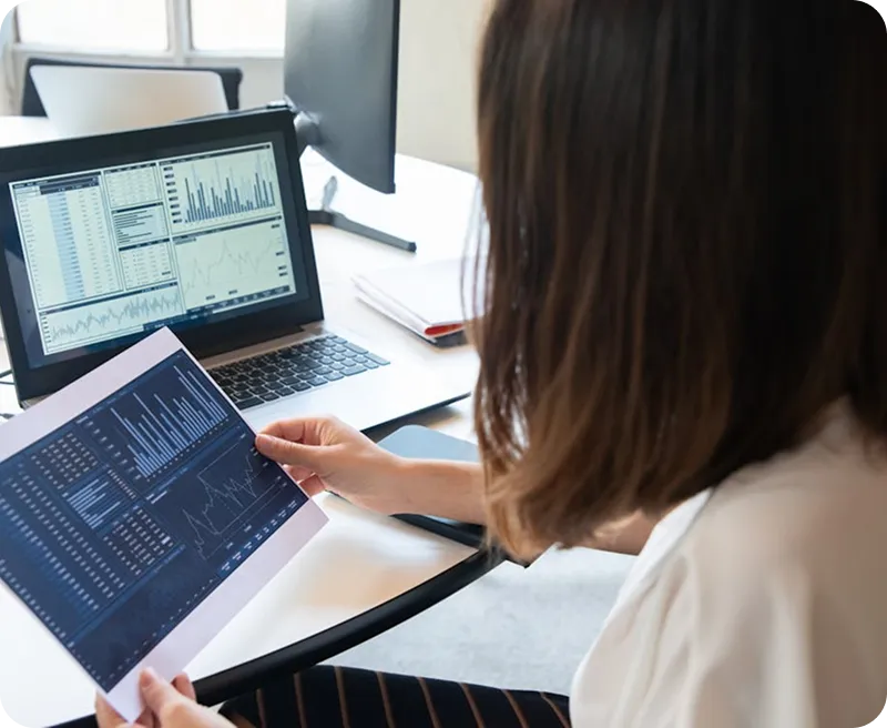 Person reviewing printed charts and graphs in front of a laptop displaying financial data on a desk.