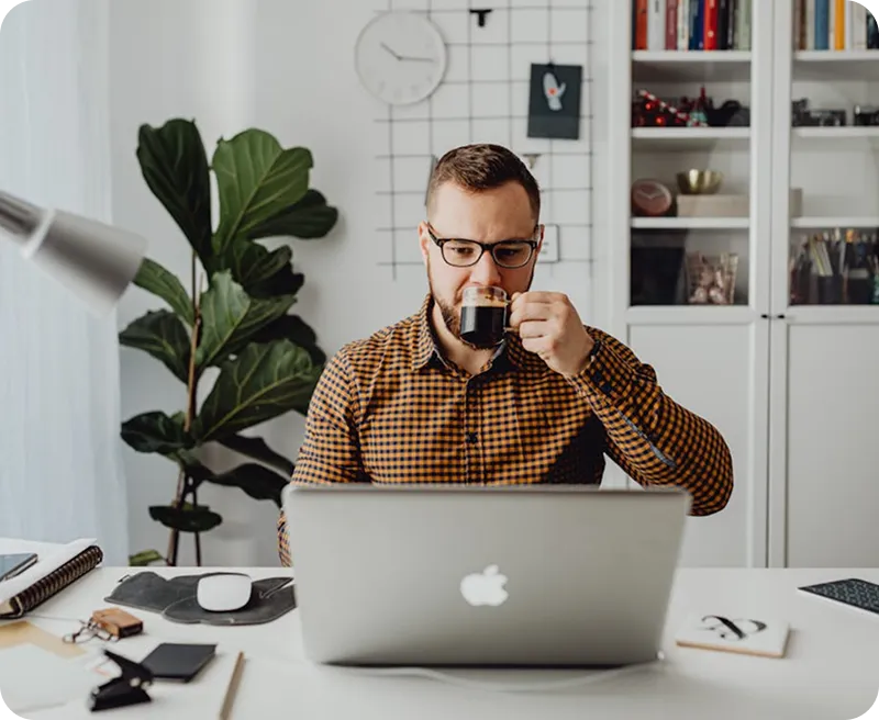 Man in glasses drinking coffee while working on a laptop at a desk in a modern home office.