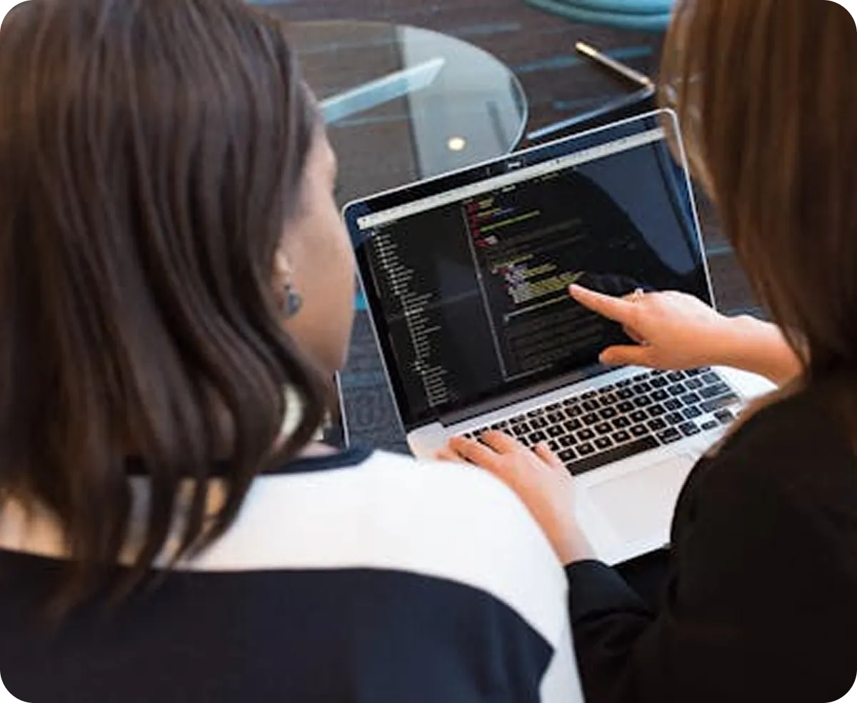 Two women working together on a laptop displaying programming code.