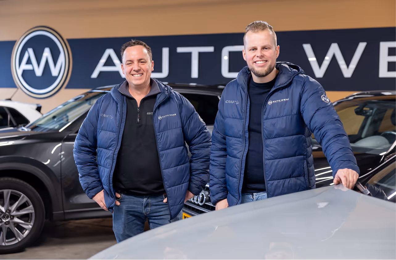 Twee medewerkers van Auto Wens staan in de showroom bij het logo van het bedrijf